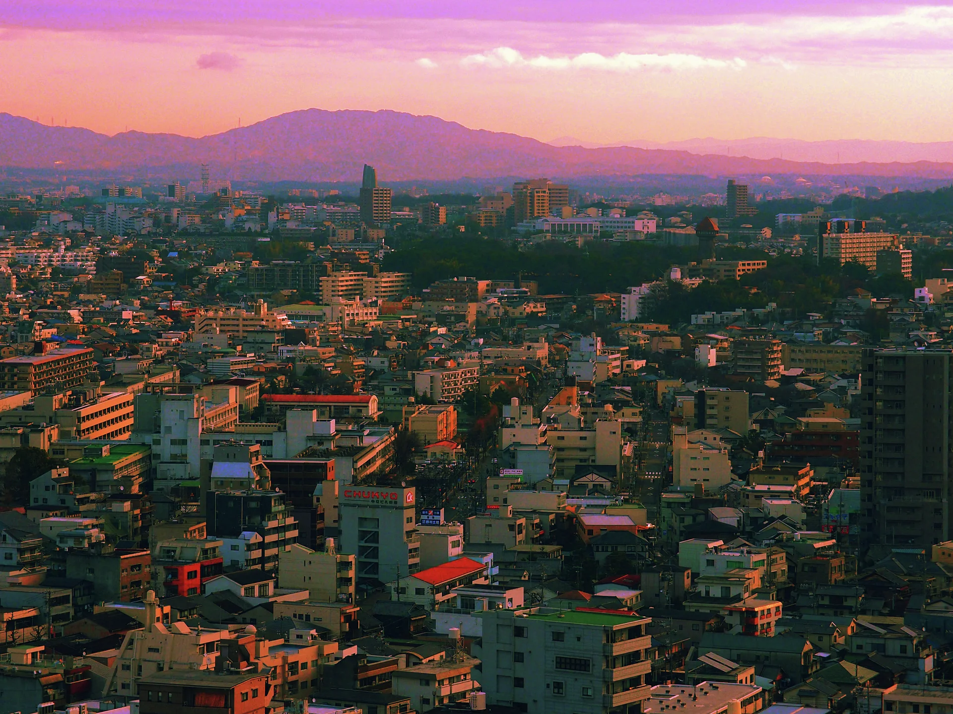 Dense city rooftops extend toward layered distant hills beneath a pink and orange sky.