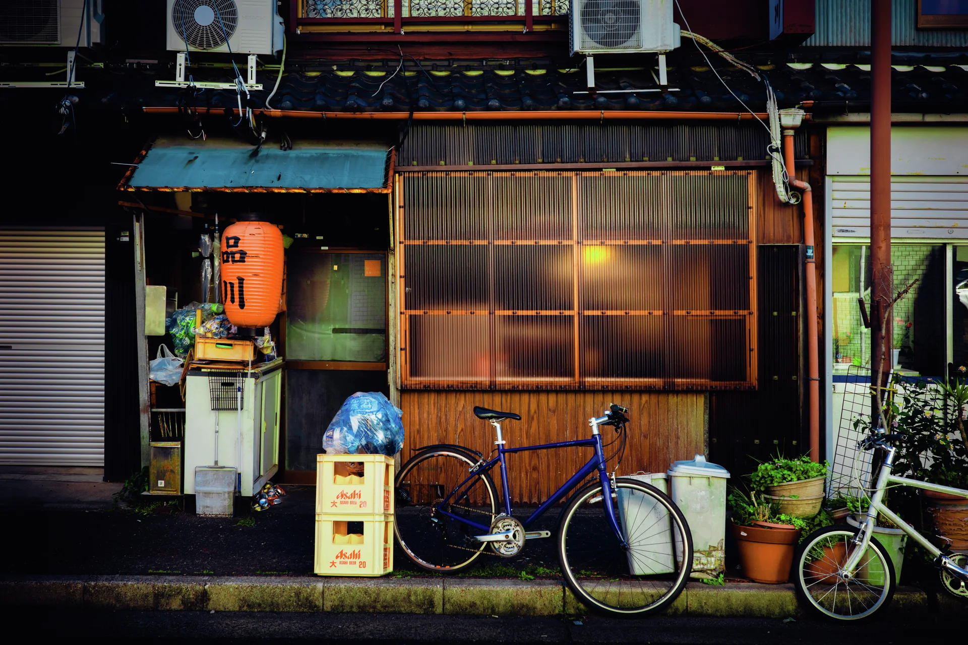 Bicycles and potted plants stand outside a wooden storefront with a glowing window and orange lantern.