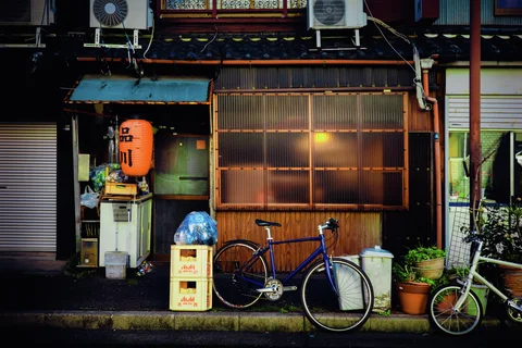 Bicycles and potted plants stand outside a wooden storefront with a glowing window and orange lantern.