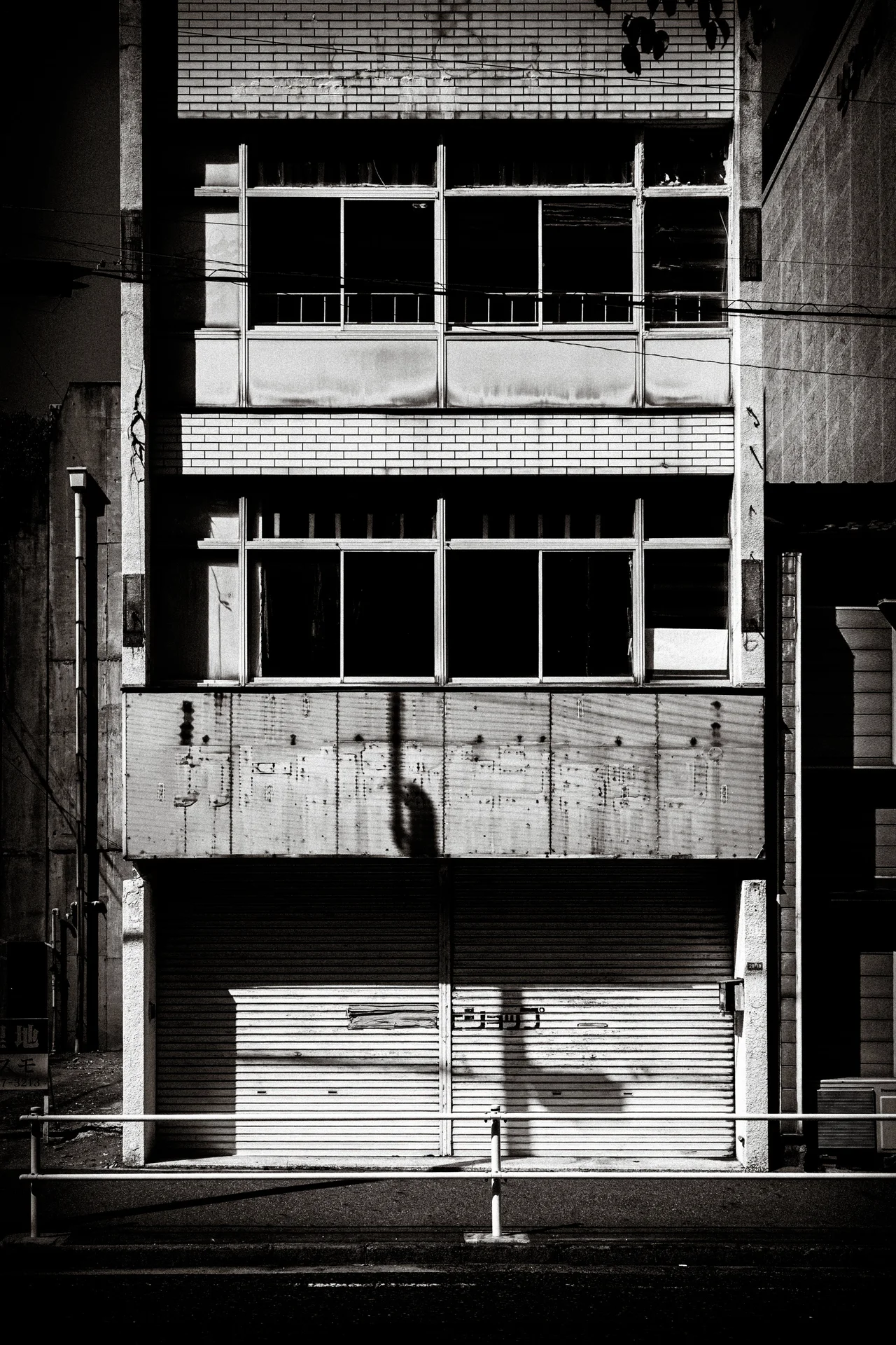 A weathered concrete facade has broad dark windows above a closed shutter in black and white.