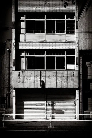 A weathered concrete facade has broad dark windows above a closed shutter in black and white.
