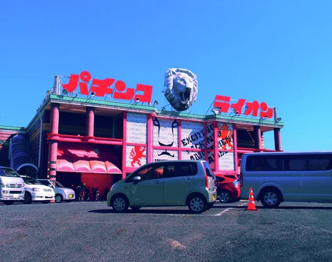 A giant lion head tops a pink commercial building with red Japanese lettering above parked cars and vans.