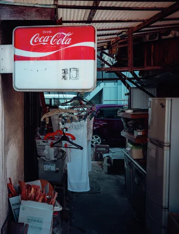 A Coca-Cola sign hangs over a narrow cluttered passage containing clothes hangers, umbrellas, and pale garments.