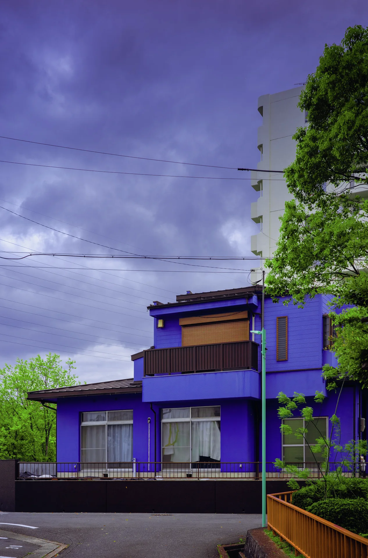 A vivid purple low building stands beneath heavy clouds beside green trees and an orange fence.