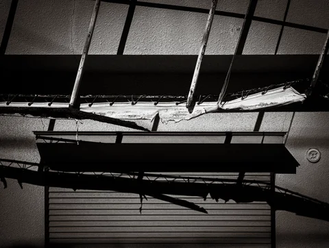 Broken canopy panels hang above a closed shutter with angular shadows in a black and white view.