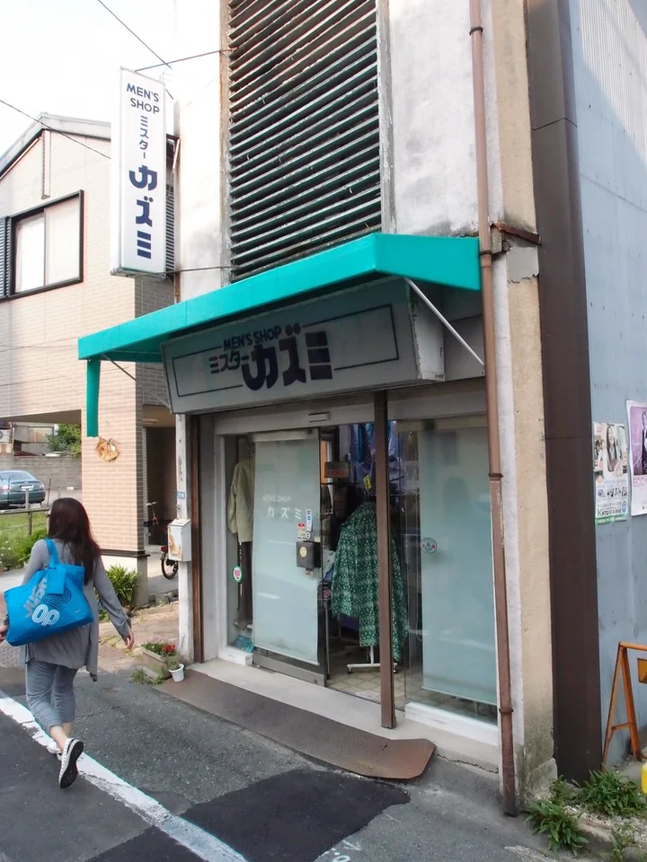 A pedestrian carrying a bright blue bag passes a narrow shop beneath a turquoise awning.