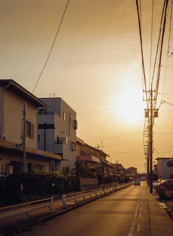 Low sunlight illuminates a quiet narrow street beneath an orange sky crossed by dense overhead wires.