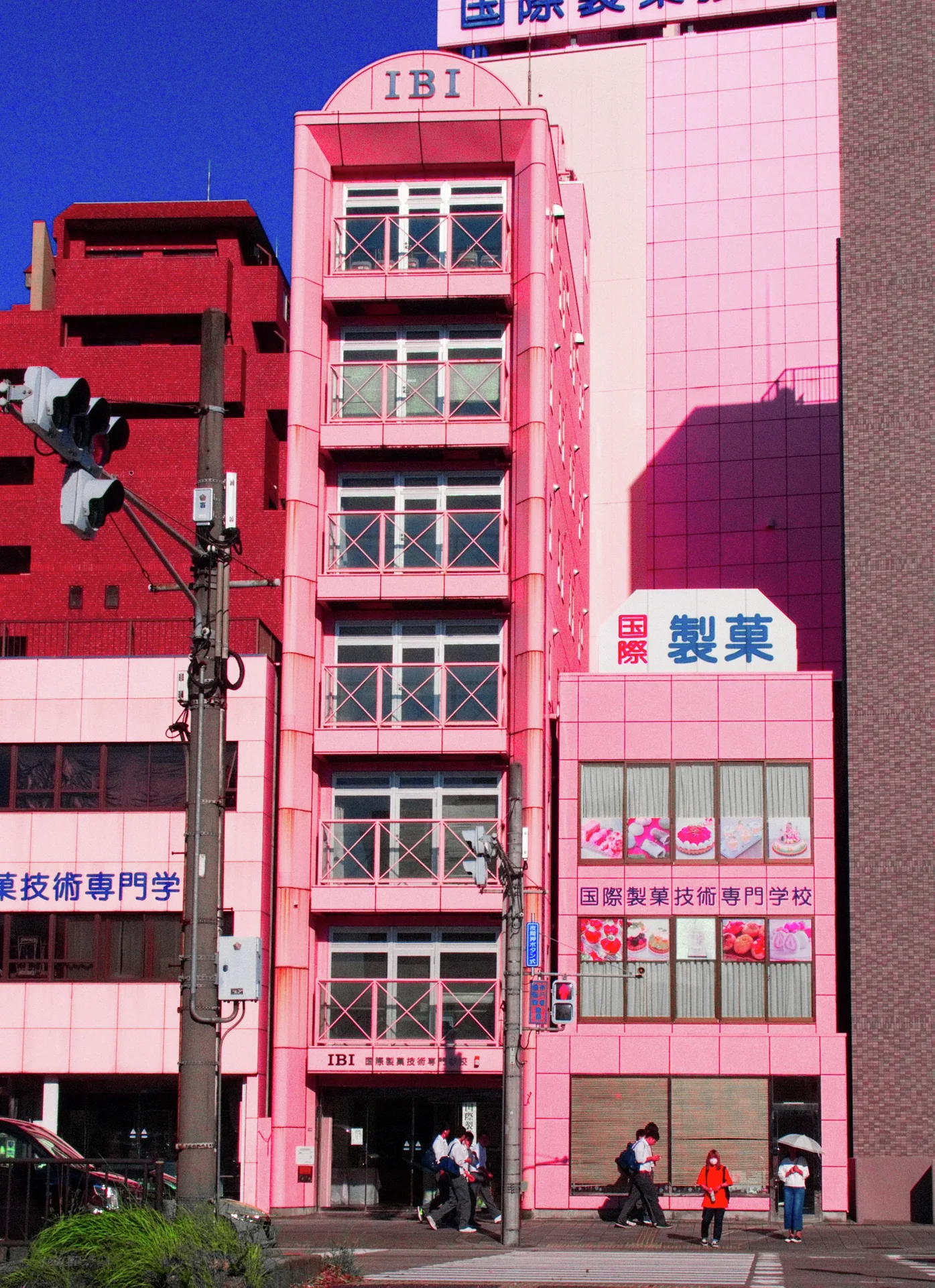 A narrow pink building has stacked windows and exterior pipes beside a larger pink facade.