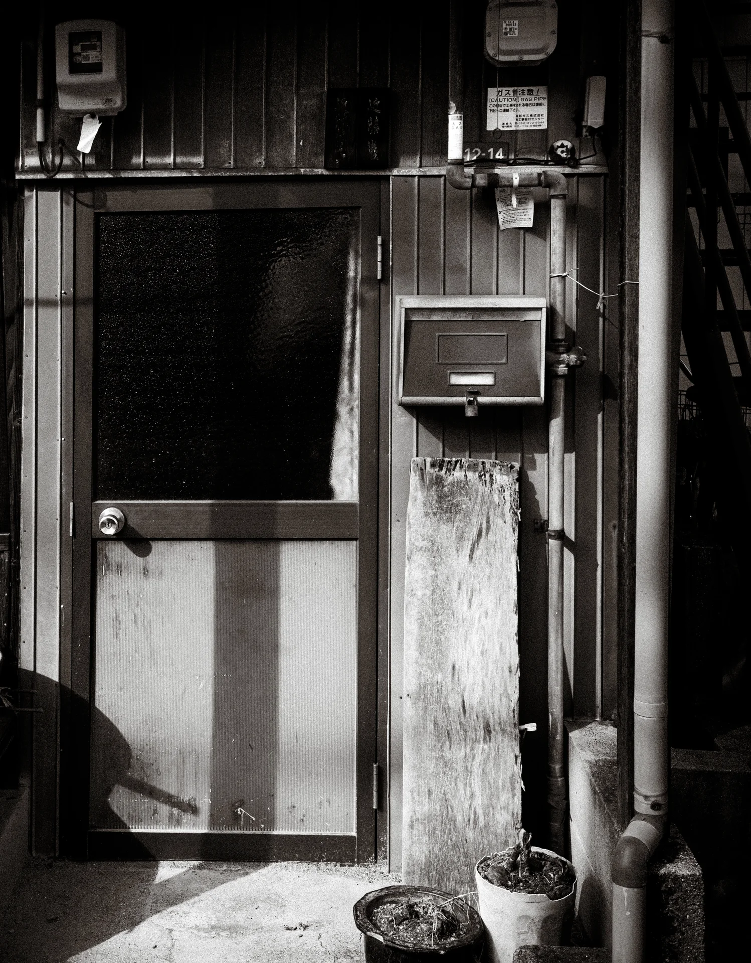A weathered doorway is bordered by a mailbox, utility meters, pipes, and small pots in black and white.