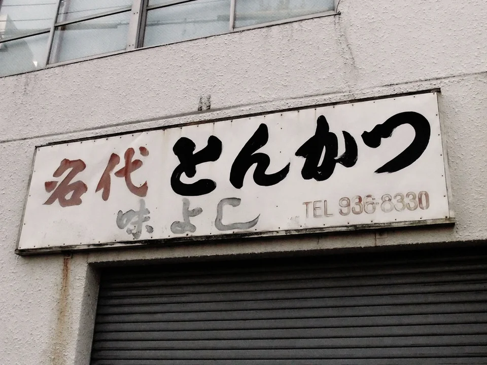 Bold dark characters fill a weathered white rectangular sign above a closed gray storefront shutter.