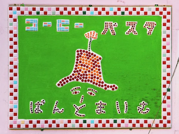 A dotted cartoon face beneath a tall hat decorates a green mosaic sign framed by multicolored square tiles.