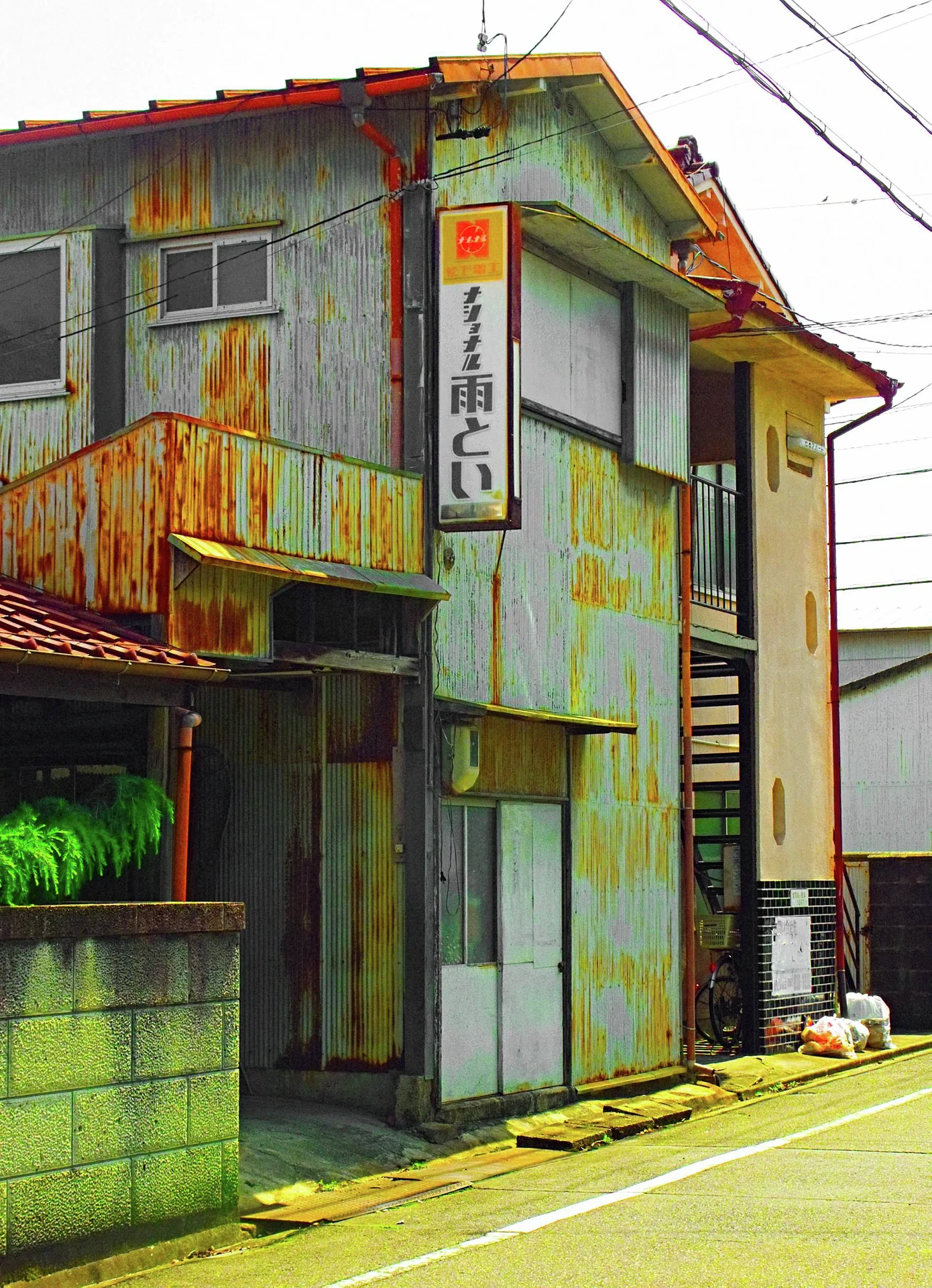Rust streaks cover an aging corrugated building with an outdoor staircase and a vertical Japanese sign.