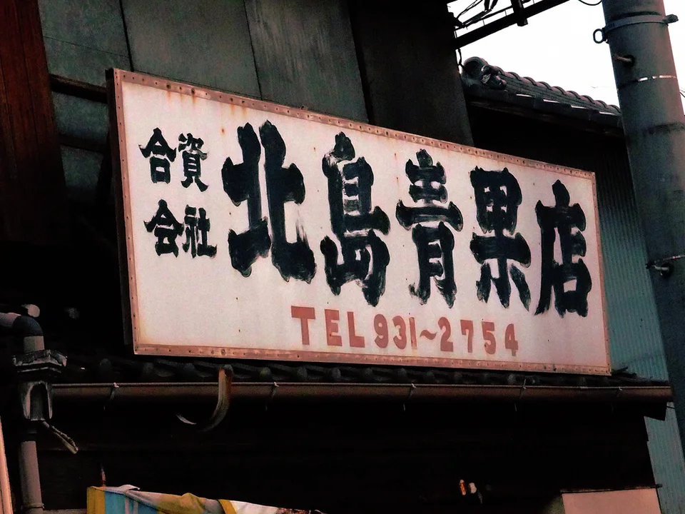 Bold black Japanese lettering fills a weathered white storefront sign above faded red telephone numbers.