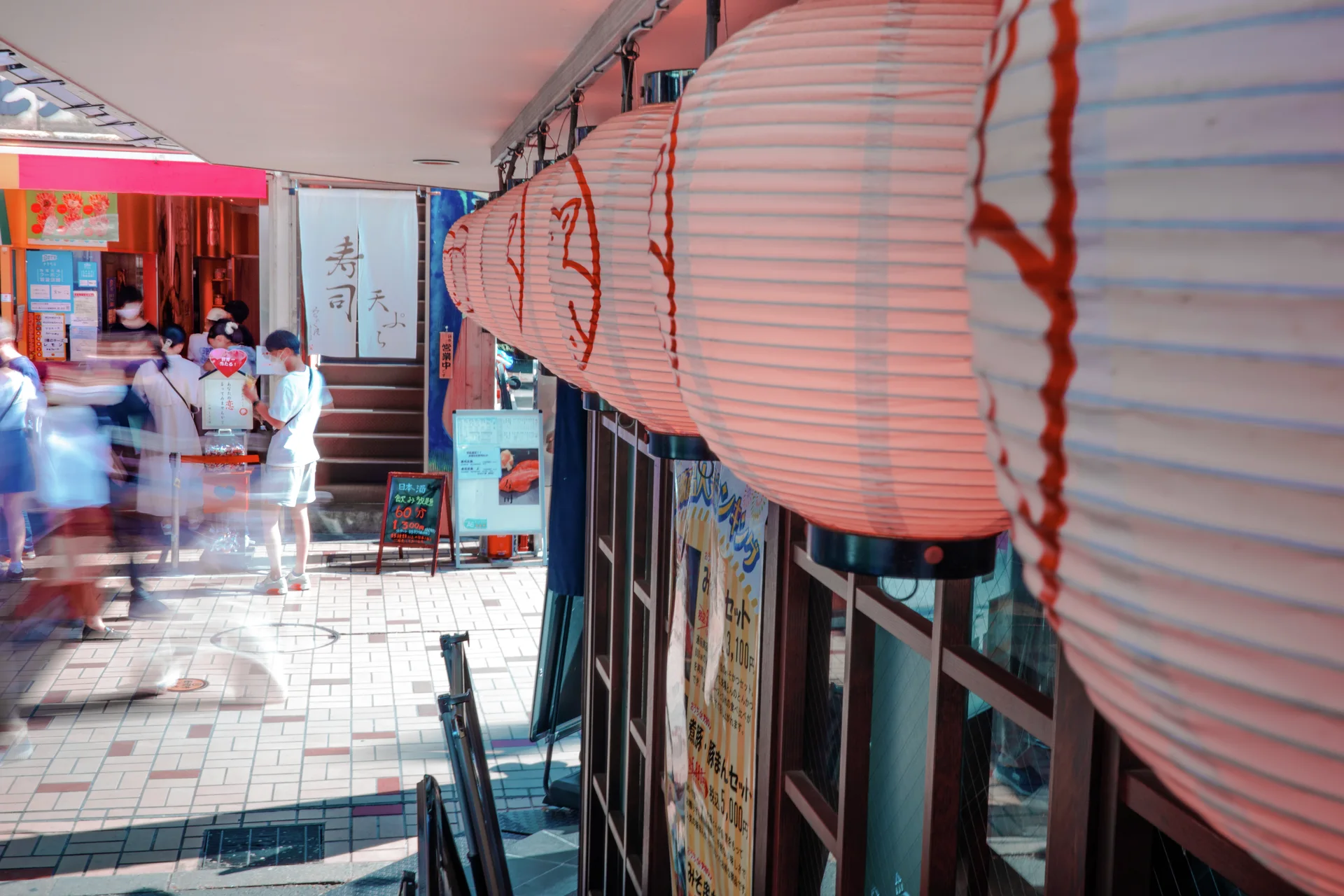 A row of large white paper lanterns hangs beside a covered shopping walkway with blurred pedestrians.