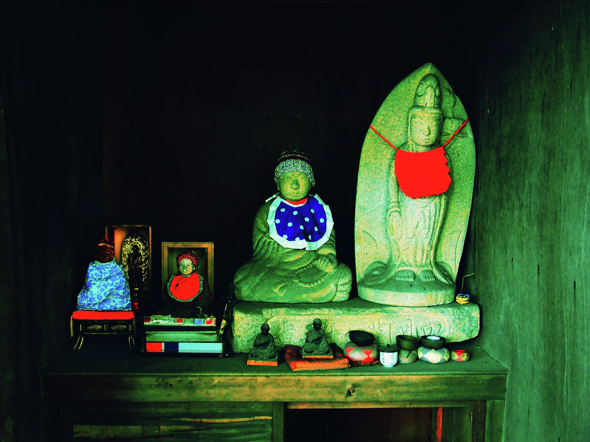 Stone religious figures wearing red and blue cloths sit on a dark shelf with small offerings.