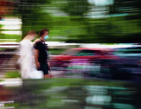 Two masked pedestrians and red cars appear against horizontally blurred greenery and street reflections.