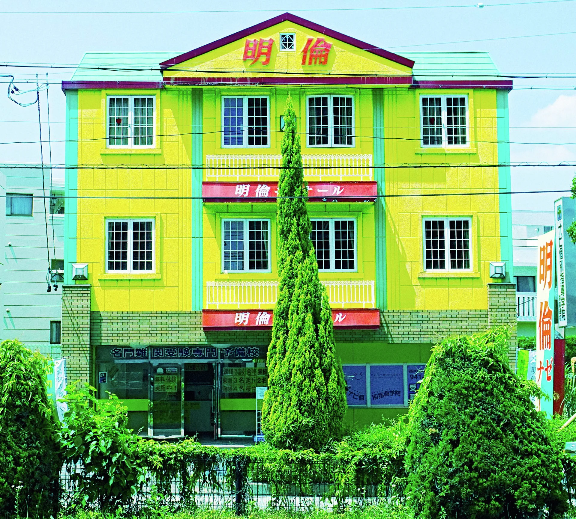 A bright yellow building with green trim and repeated grid windows rises behind neatly shaped shrubs.