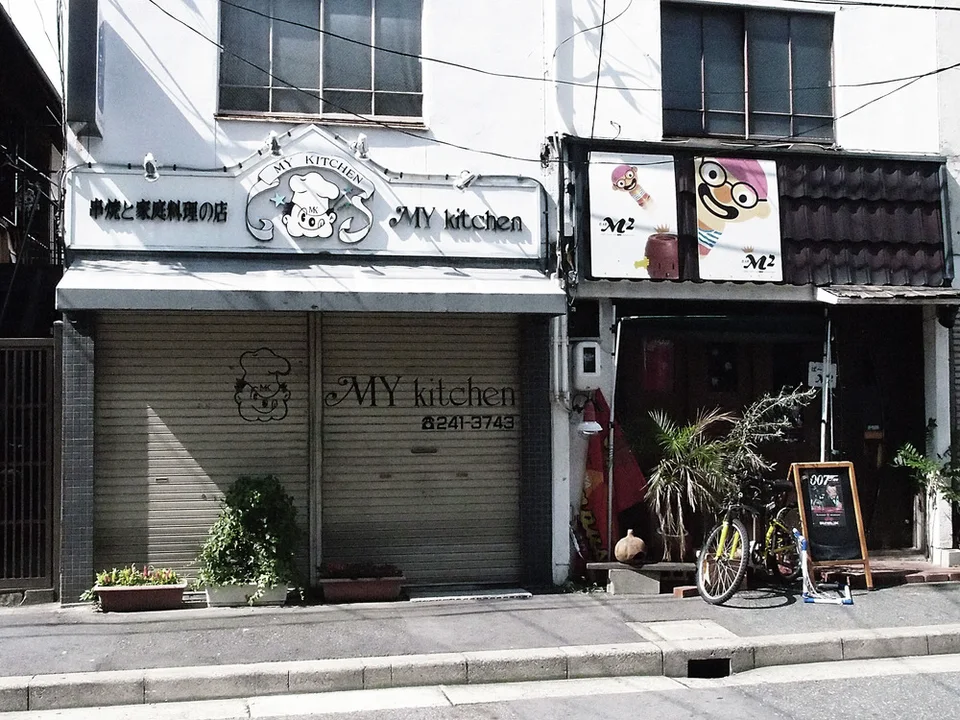 Two small adjacent storefronts have illustrated signs, a closed shutter, and a bicycle beside plants.