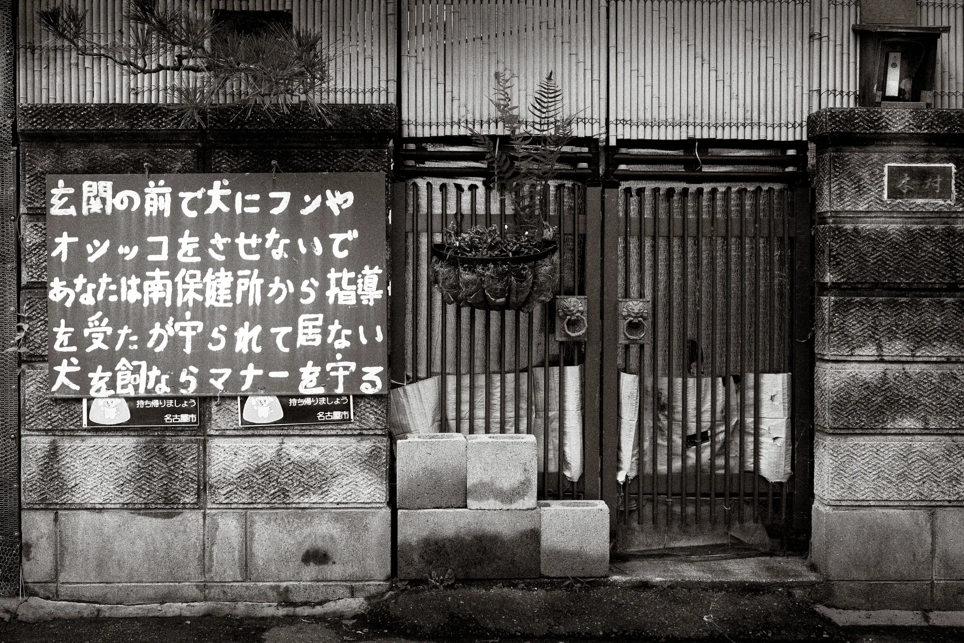 A handwritten Japanese notice hangs beside a weathered wooden gate and block wall in black and white.
