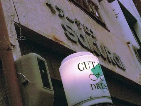 A pale cylindrical salon sign projects below raised lettering on a stained wall beside an outdoor air conditioner.
