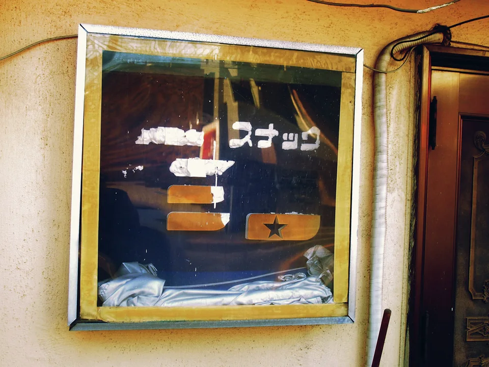 A framed dark shop sign with faded lettering hangs on a yellow wall beside exposed cables.