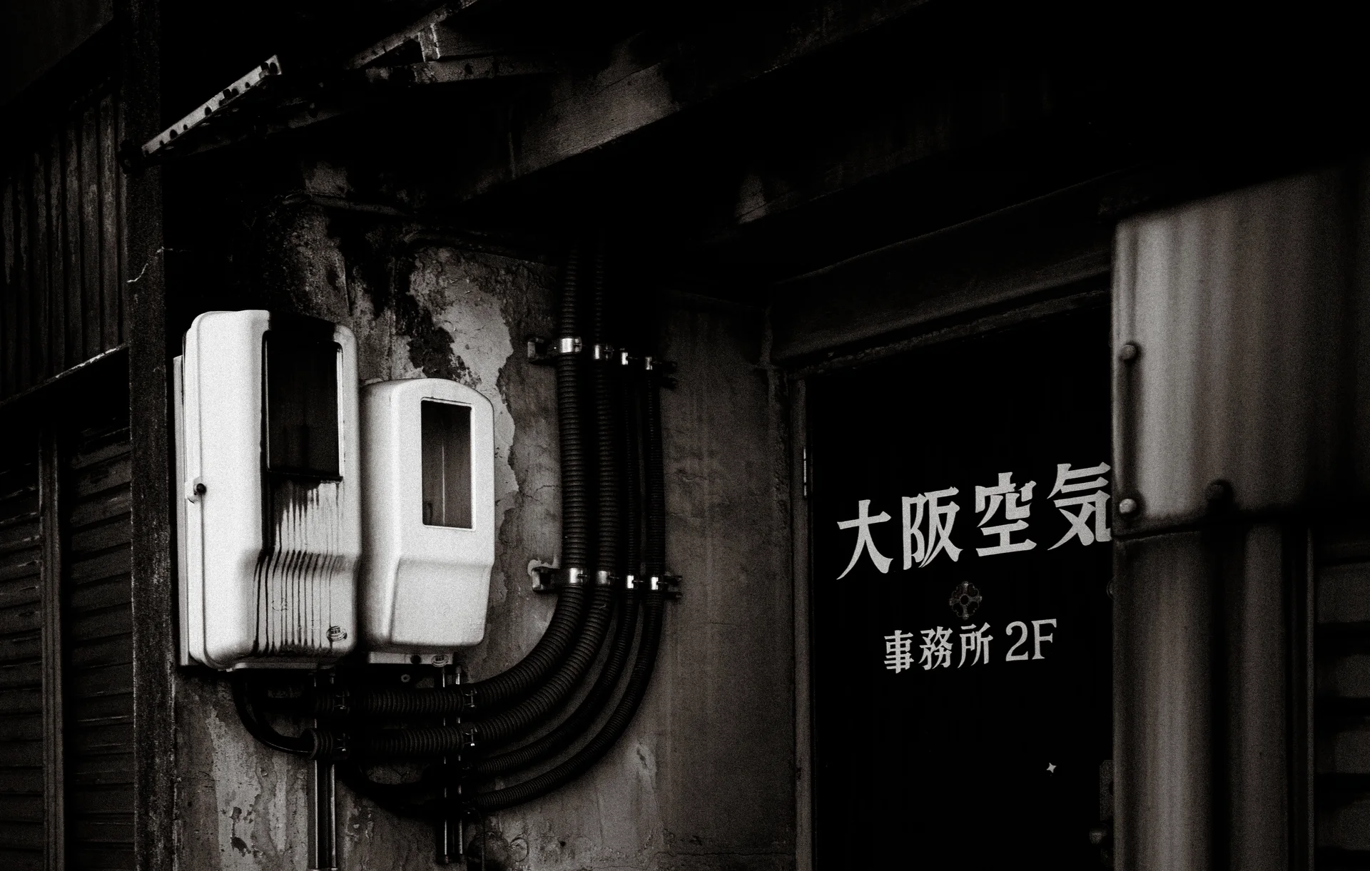 Two pale electrical boxes and curved black conduits sit beside a dark lettered door.