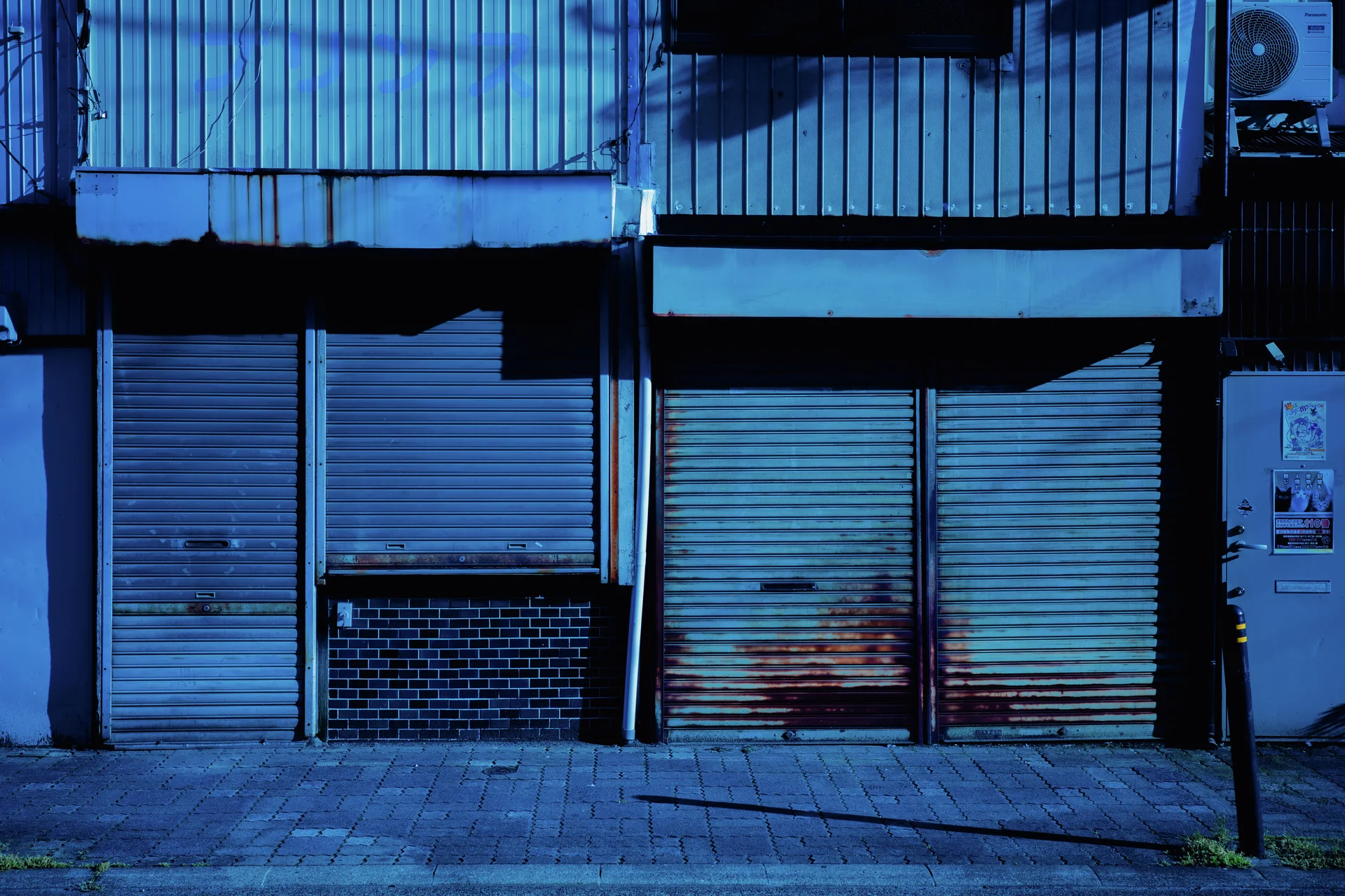 Closed metal shutters and corrugated walls appear in deep blue tones with rusty patches near the pavement.