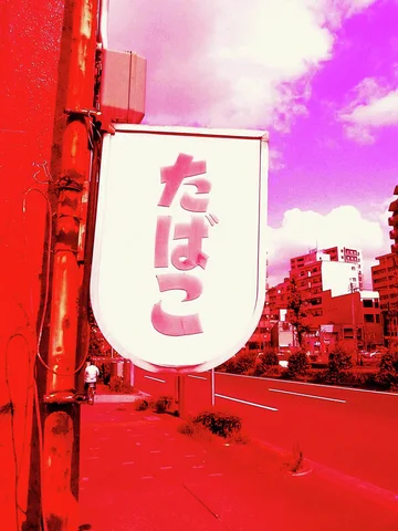 A rounded white tobacco sign with pink Japanese lettering projects beside a vivid red street under pink clouds.