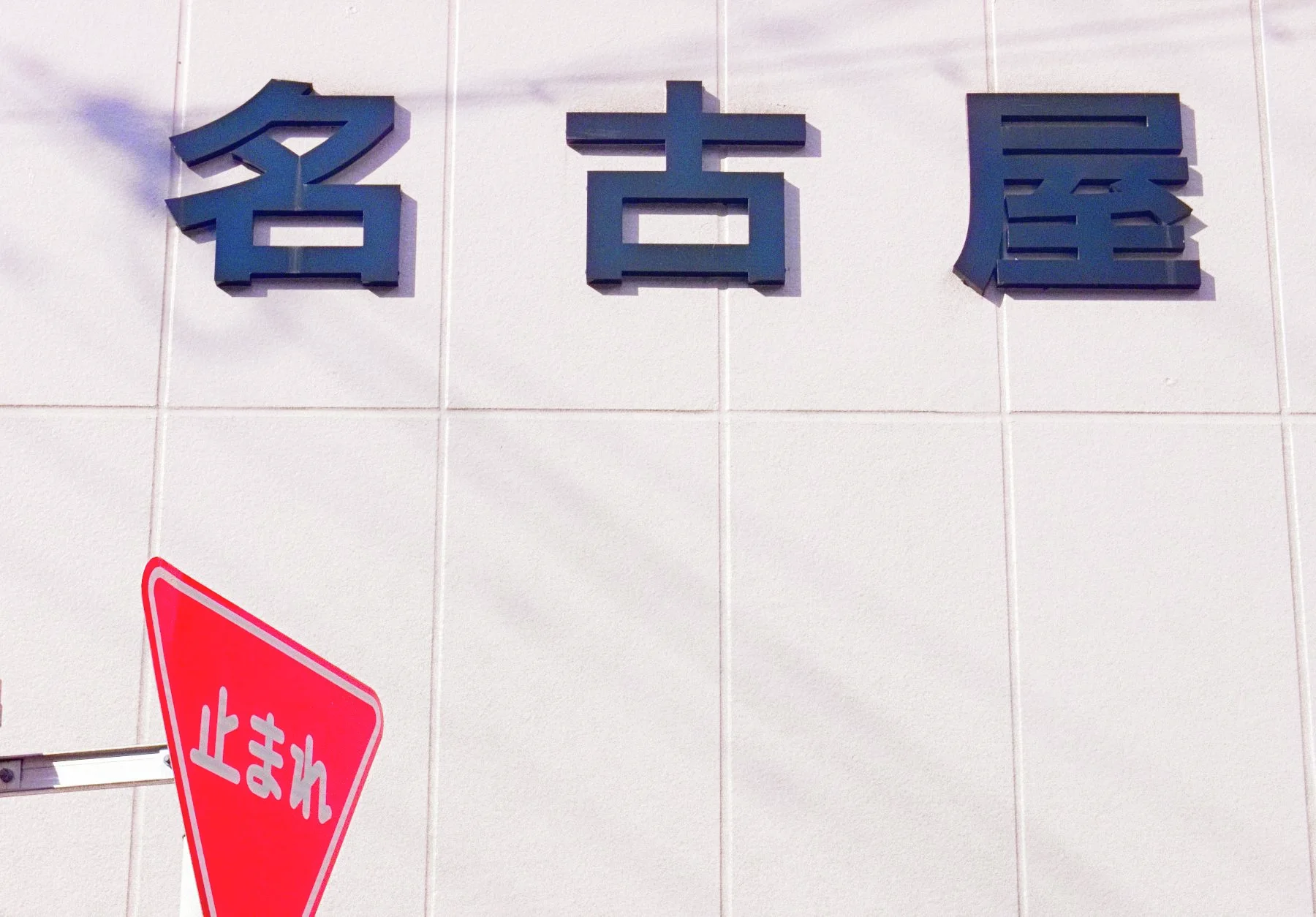 Three large dark Japanese characters project from a pale tiled wall above a red triangular street sign.