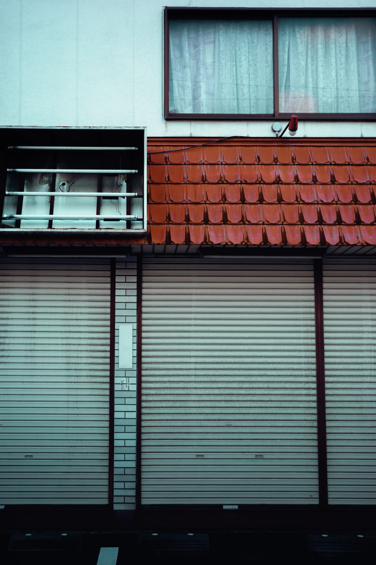 Closed pale shutters line a building beneath a red tiled awning and a curtained upstairs window.