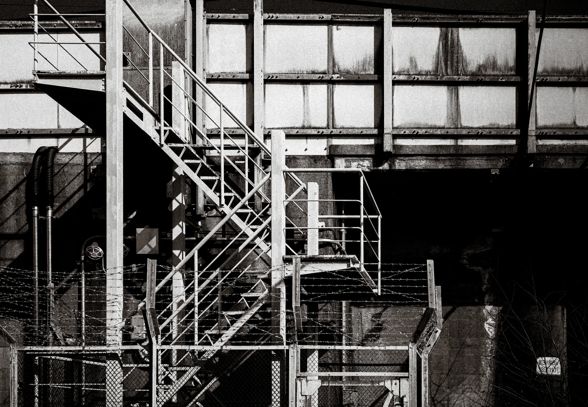 An exterior metal staircase zigzags across a weathered industrial wall behind fencing in monochrome.