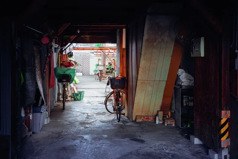 A dark workshop passage contains leaning boards and a bicycle, opening onto a brighter fenced yard.