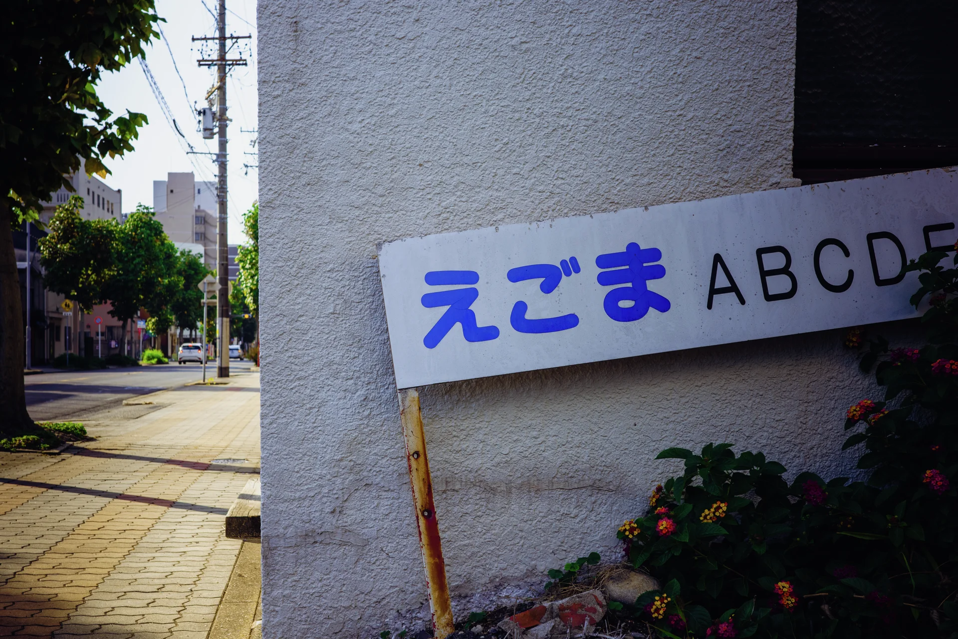 A white sign with blue Japanese lettering and black alphabet letters rests against a textured wall.