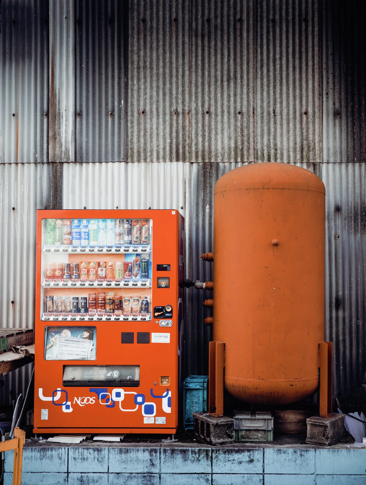 An orange drinks vending machine stands beside a tall orange tank against a corrugated metal wall.