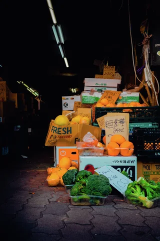 Boxes of orange fruit and leafy vegetables crowd the entrance of a dark shop beneath fluorescent lights.