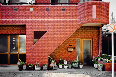 Potted flowers line a red tiled facade beneath an angular enclosed exterior staircase.