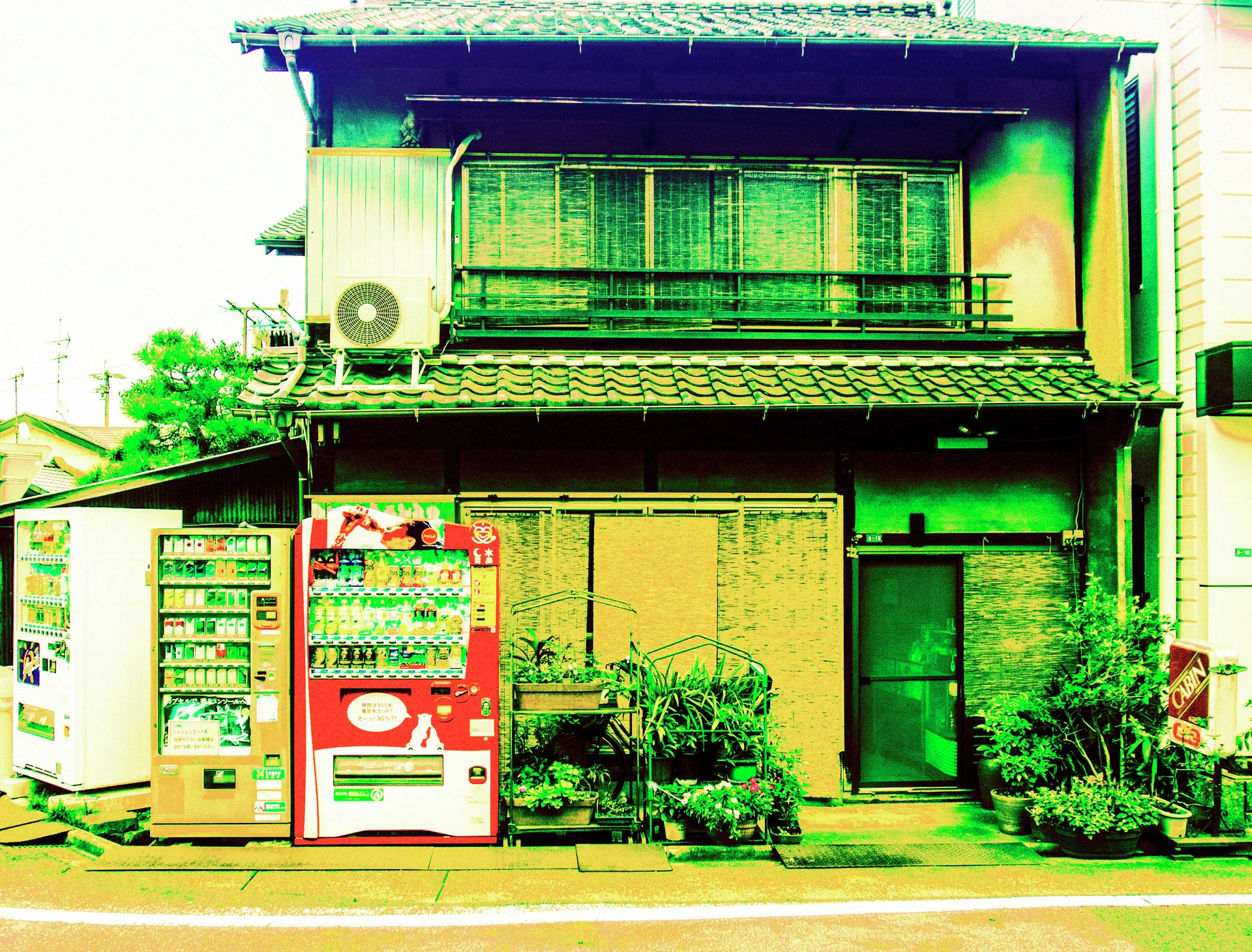 Three vending machines and potted plants stand outside a weathered tiled-roof house with screened upstairs windows.