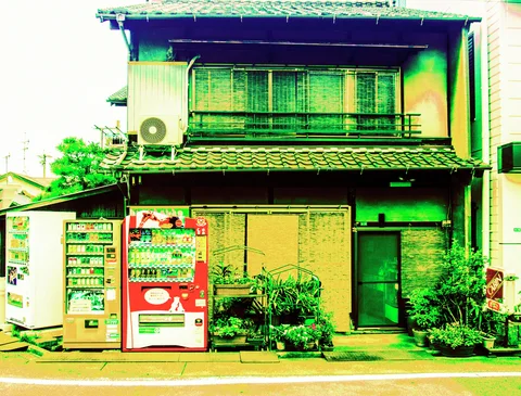 Three vending machines and potted plants stand outside a weathered tiled-roof house with screened upstairs windows.