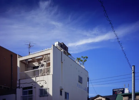 A white building with open balconies rises beneath wispy clouds and overhead wires against blue sky.