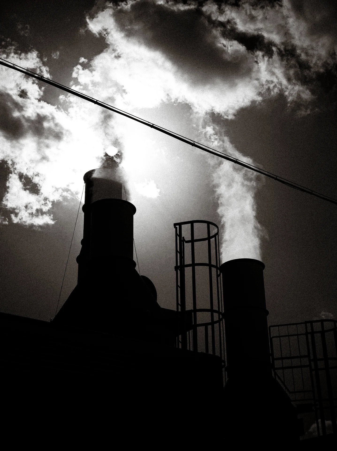 Industrial stacks silhouette against a bright cloud opening crossed by a diagonal overhead wire.