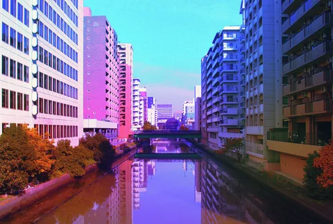 Tall colorful buildings and trees line a calm canal reflecting the sky beneath a distant bridge.