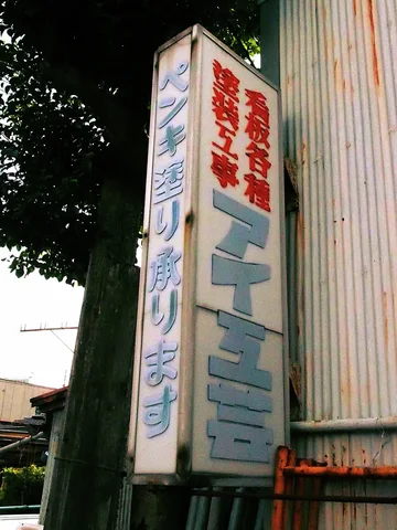 A weathered projecting sign bears red and pale blue Japanese lettering beside corrugated siding and leafy trees.