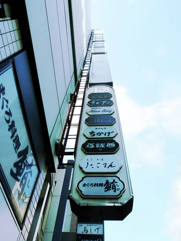 Stacked rounded business labels climb a narrow projecting directory against pale blue sky beside a building wall.