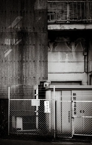 A chain-link fence borders a stained industrial building beneath a projecting balcony in black and white.