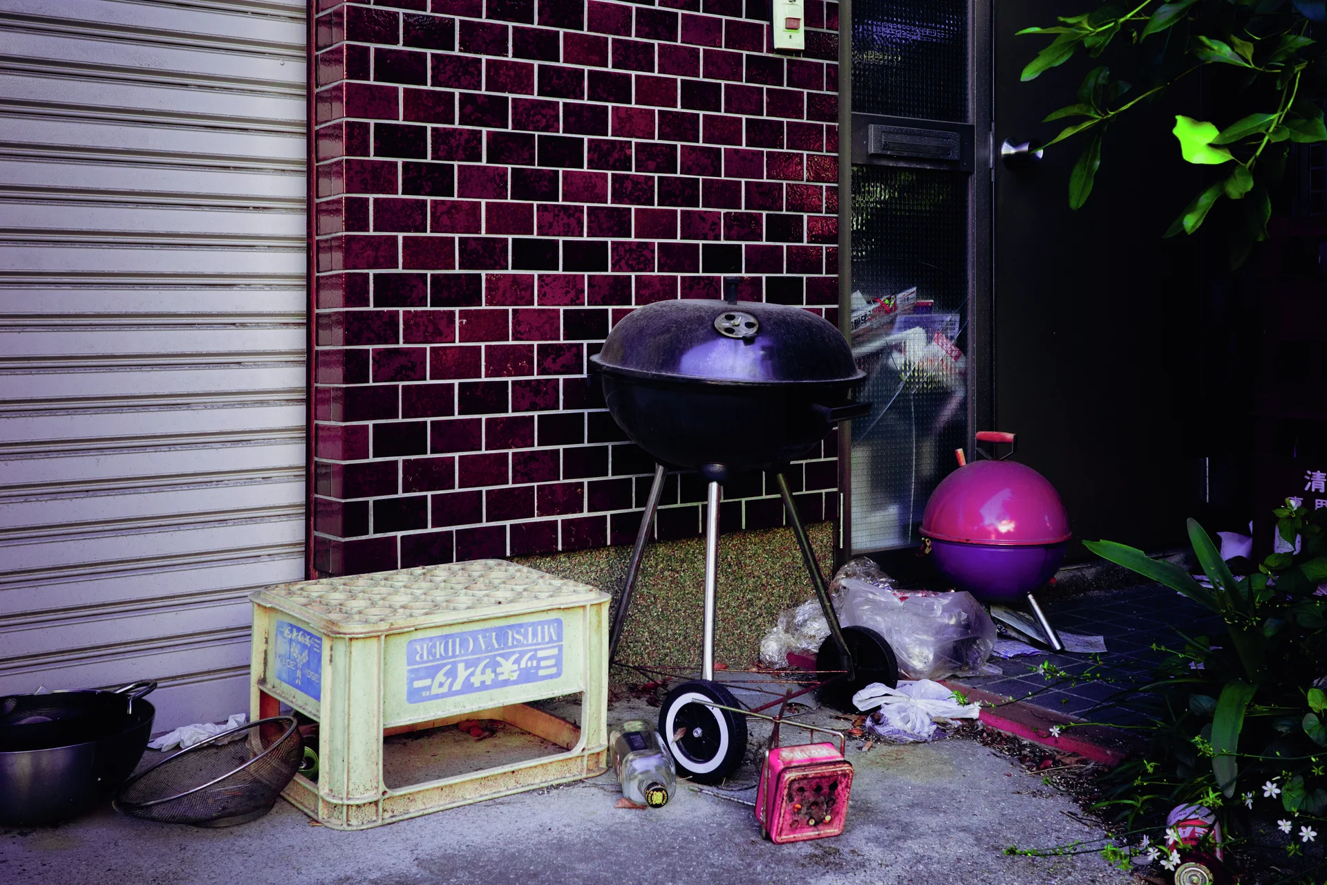 A black kettle grill and smaller pink grill stand among scattered objects beside dark red tiles.