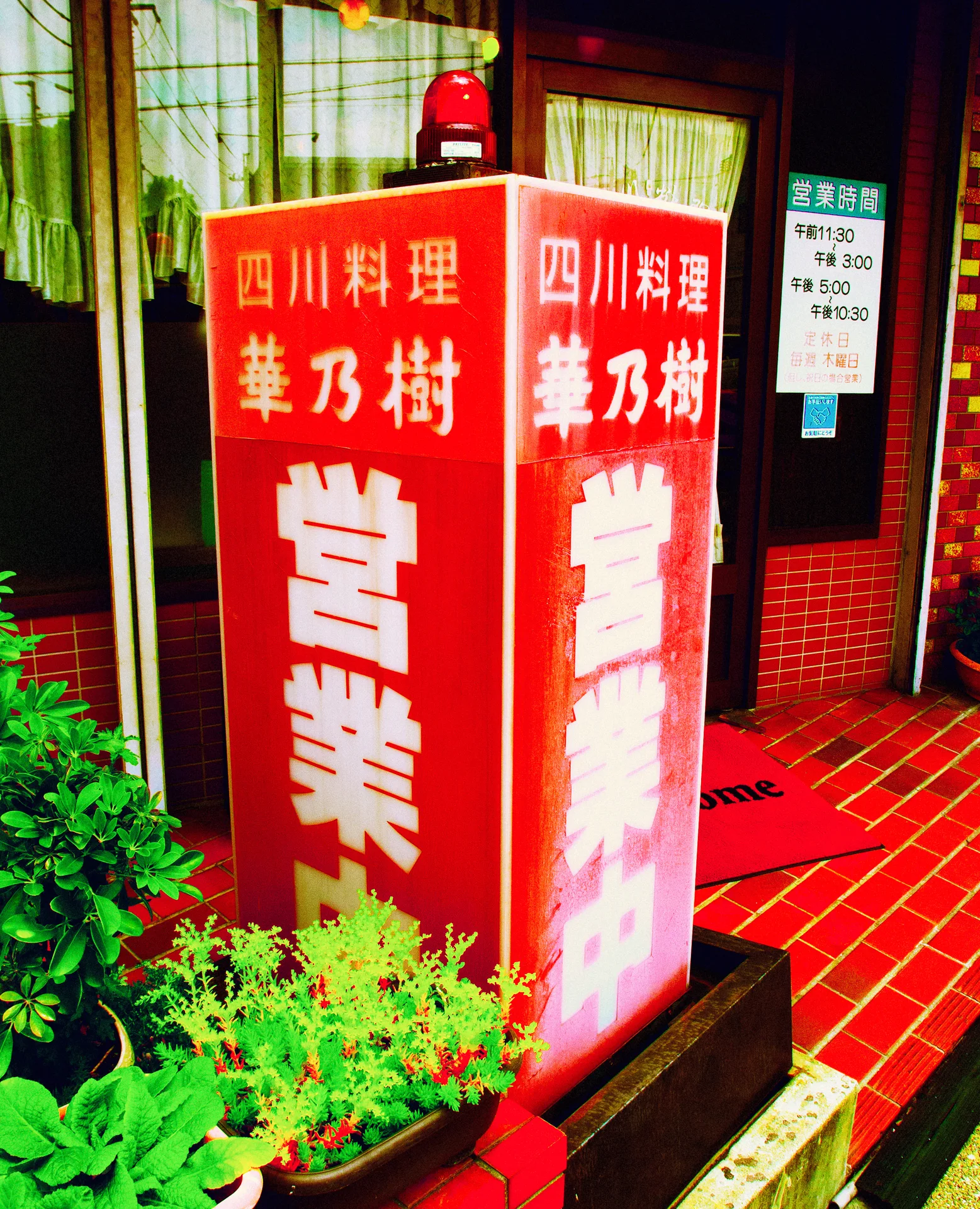 A tall red illuminated restaurant sign stands beside potted greenery and a red tiled entrance.