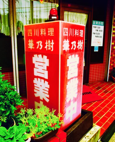 A tall red illuminated restaurant sign stands beside potted greenery and a red tiled entrance.