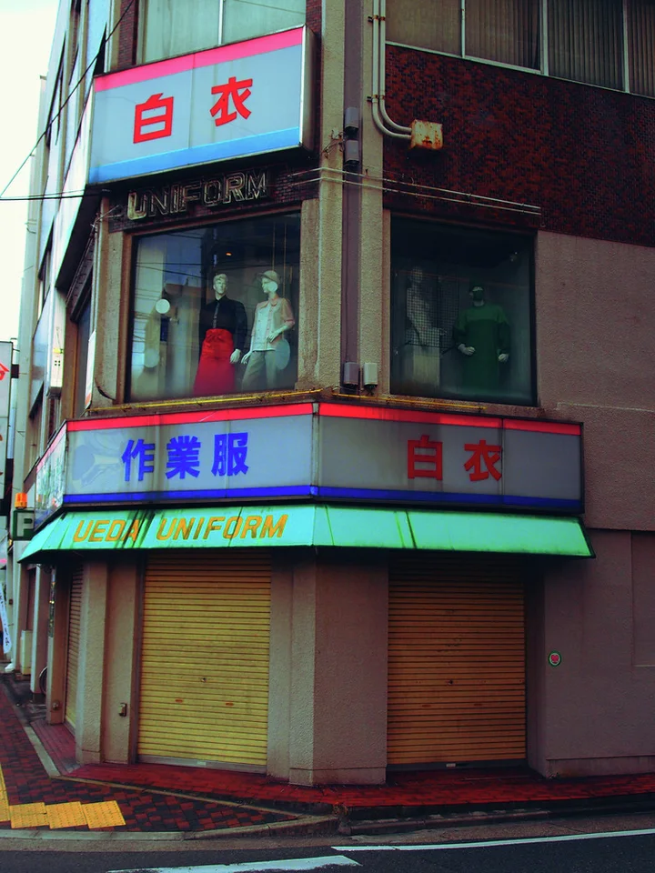 A corner uniform shop has closed yellow shutters beneath blue signs and an upstairs mannequin display.