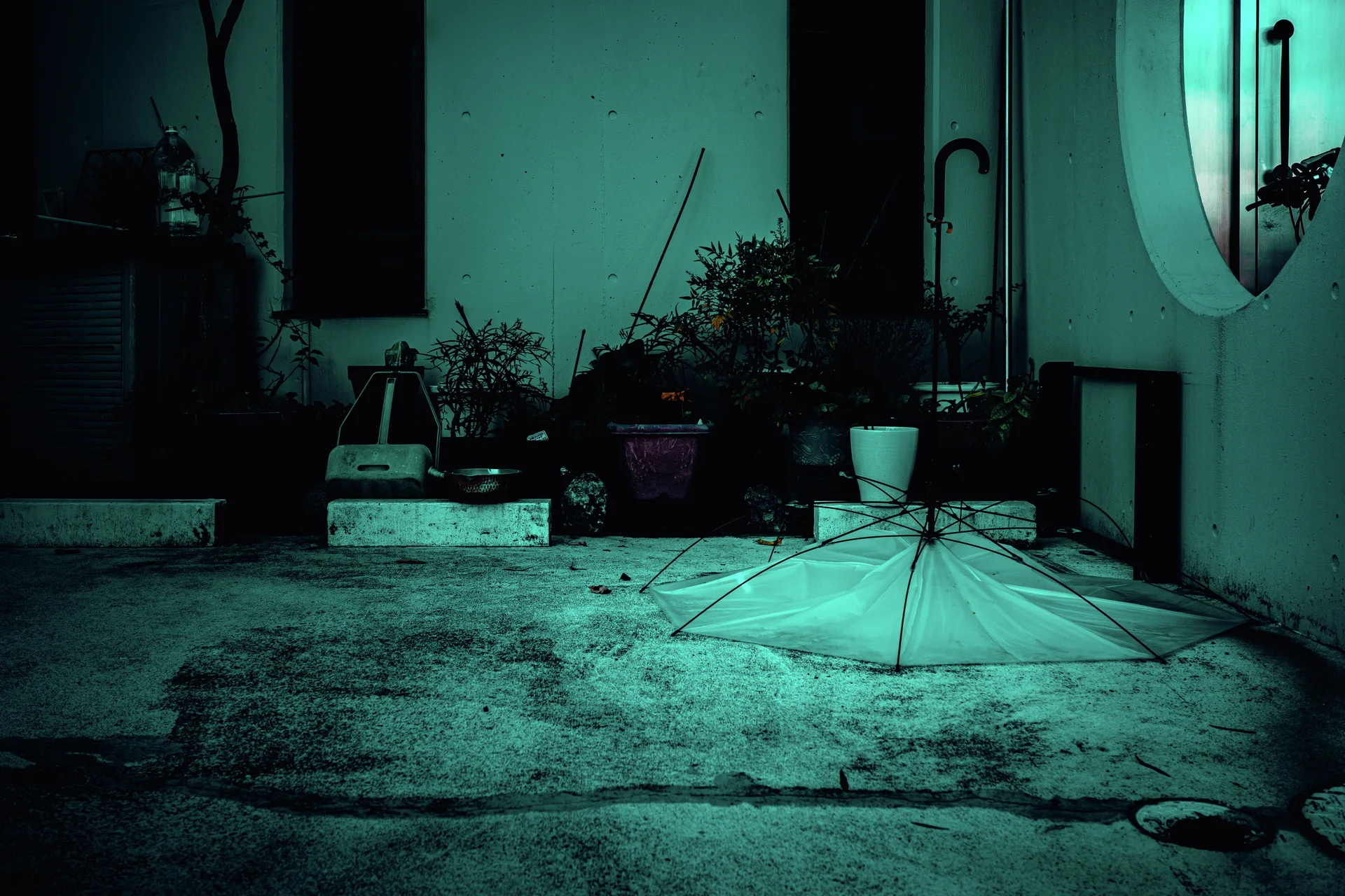 An open pale umbrella rests on turquoise-toned concrete beside potted plants and exterior pipes.