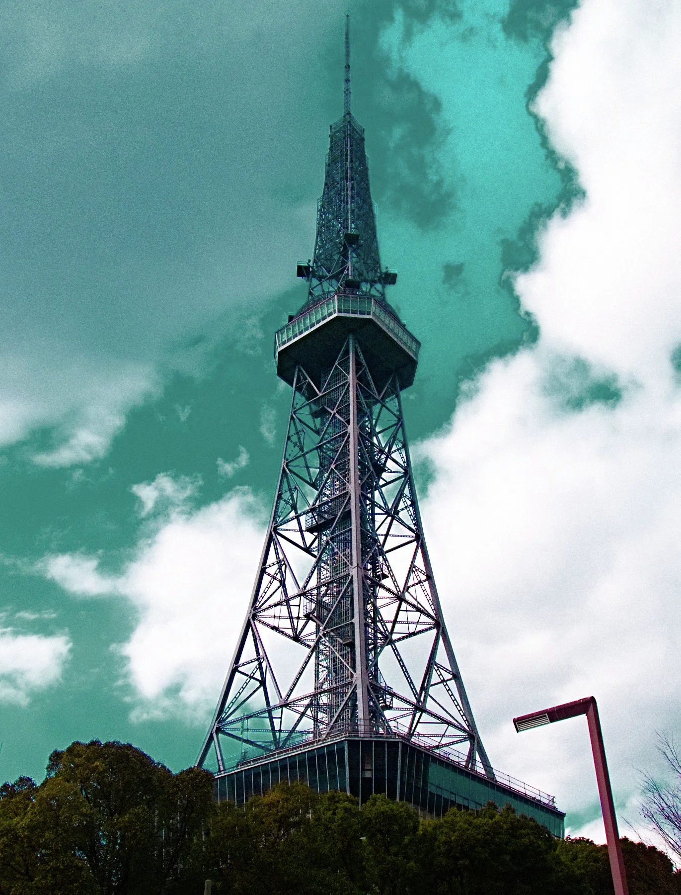 A steel lattice tower with a broad observation section rises above treetops beneath turquoise sky and white clouds.
